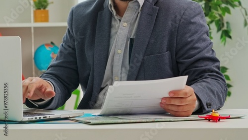 A man in a suit sits in a travel agency office and enters data from a document into a laptop.