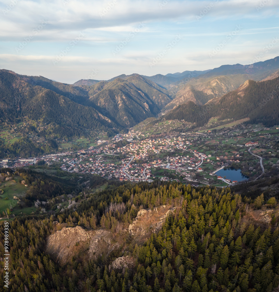 Obraz premium A spring mountain valley at sunrise with rich vegetation and spruce dark dense forests and a small picturesque town of Smolyan nestled between the slopes, Rhodopi Mountains, Bulgaria.