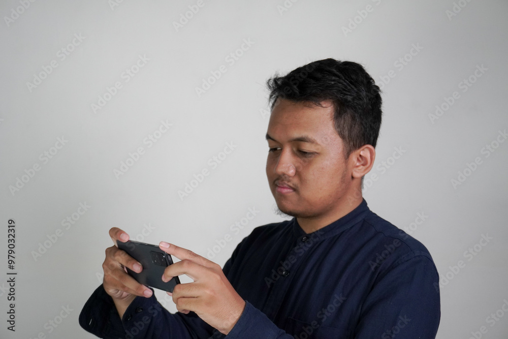 A Southeast Asian man wearing a dark blue shirt is using his cell phone, tilting the phone to watch something on his cell phone. A Southeast Asian man wearing a dark blue shirt is using his cell phone, tilting the phone to watch something on his cell phone.