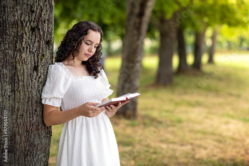 A woman standing in a meadow under a tree in the summer. Young girl in white dress relaxing studying and reading book in nature on vacation