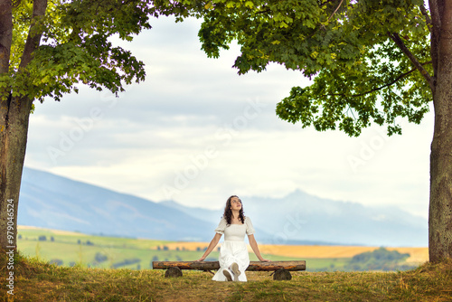 Young woman sitting on a wooden bench under the trees looking at the leaves of the trees in a white dress. Girl in nature with a view of the mountains in the background