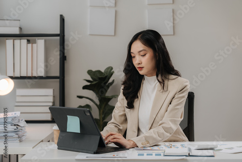 Focused Professional: A young businesswoman in a stylish blazer, engrossed in work on her tablet at her desk, surrounded by paperwork and a plant, radiates determination and focus. 