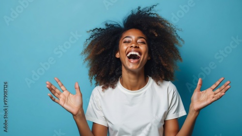 Curly optimistic woman raises palms from joy, happy to receive an awesome present from someone, shouts loudly, dressed in casual white t-shirt, isolated on blue background. Excited Afro female