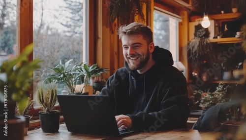 Smiling Young Man Working Remotely on Laptop in Cozy Cafe with Plants, Freelancer Lifestyle