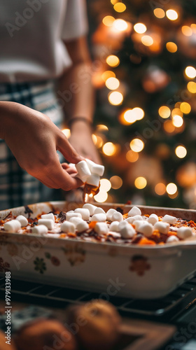 Hand placing marshmallows on sweet potato casserole with Christmas tree lights in background