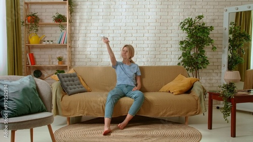 Young millennial woman sitting on the sofa in the living room feels summer heat, waving hands, turns on air conditioner. Stuffiness everyday life concept. photo