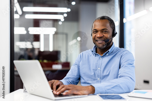 African american mature businessman working on laptop in office setting. Wearing headset for professional communication. Bright and modern workspace conveys professionalism and engagement.