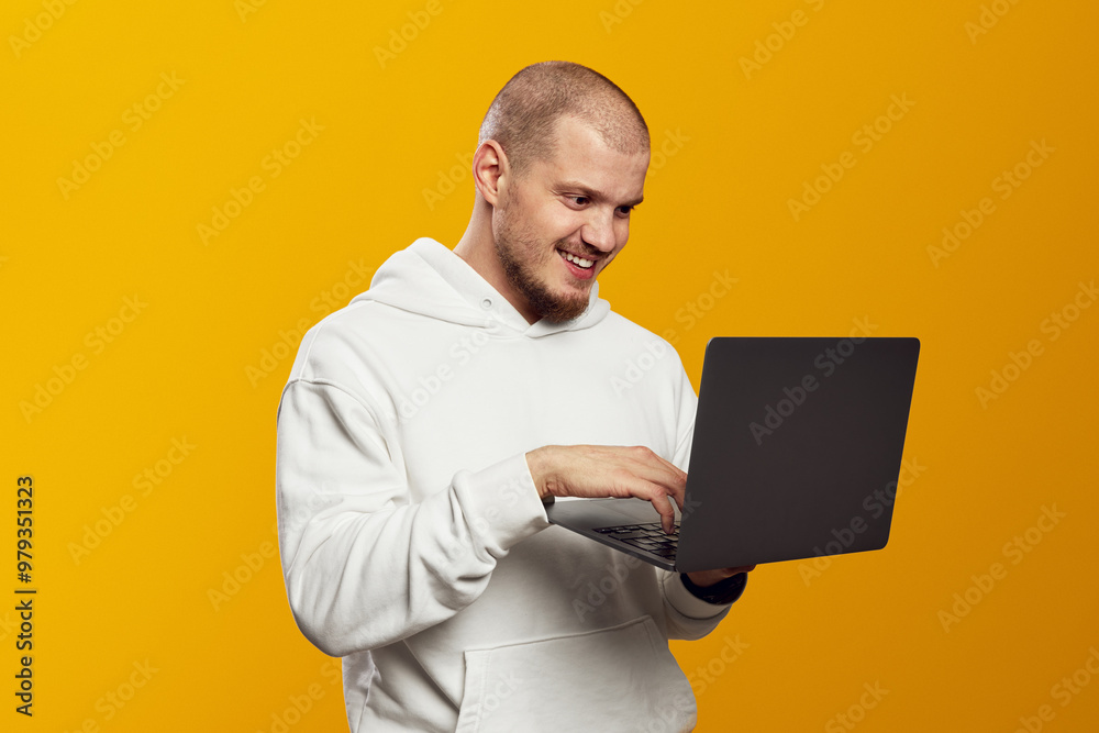 Happy adult caucasian guy in white hoodie holding laptop works remotely isolated on yellow background. Happy adult caucasian guy in white hoodie holding laptop works remotely isolated on yellow background.