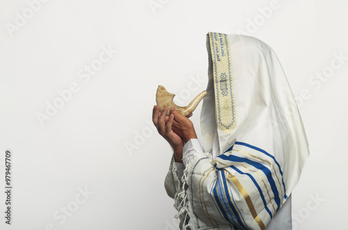 Man wearing a tallit, traditional Jewish prayer shawl with blue stripes and Hebrew text, blowing into a shofar, rams horn used in Jewish ceremonies, Rosh Hashanah. Elements of Jew religious tradition