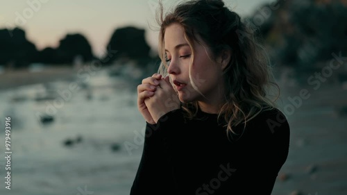 A woman stands on a chilly beach, hands clasped together as she tries to warm up. The ocean waves create a serene background, capturing a moment of solitude and reflection.