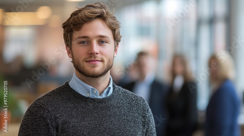 A young bearded man wearing a grey sweater and blue shirt stands confidently in an office environment with colleagues blurred in the background