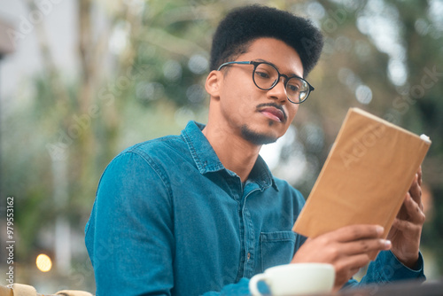 Pensive young man in casual wear deeply engaged in a book at an outdoor café. Reflective student enjoying a quiet reading moment with coffee on a peaceful day.