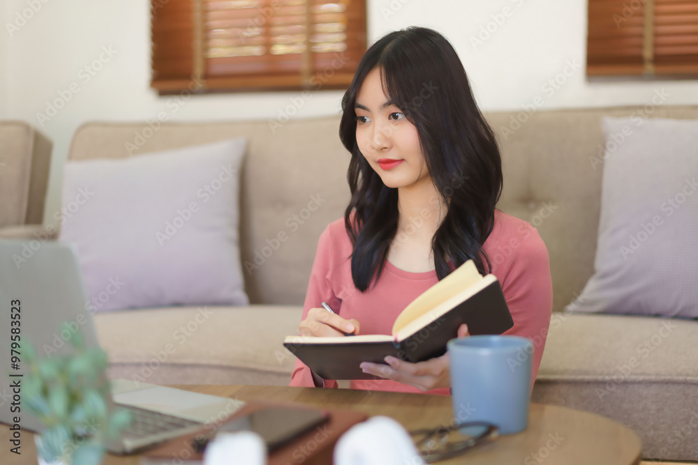 Young businesswoman thoughtful and taking notes in notebook while reading business data on laptop Young businesswoman thoughtful and taking notes in notebook while reading business data on laptop