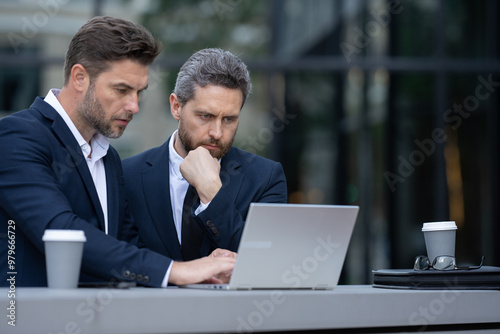 Two businessman on the street, deeply engrossed, eyes fixed on their laptop screen. Two handsome young businessmen in classic suits using laptop. Business men team using laptop outdoor. Business man