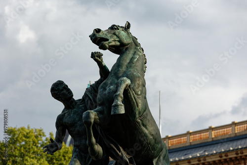 A dynamic bronze statue features a muscular man taming a powerful rearing horse against a bright sky. A striking visual inspiring concepts of strength, leadership, control and overcoming challenges.