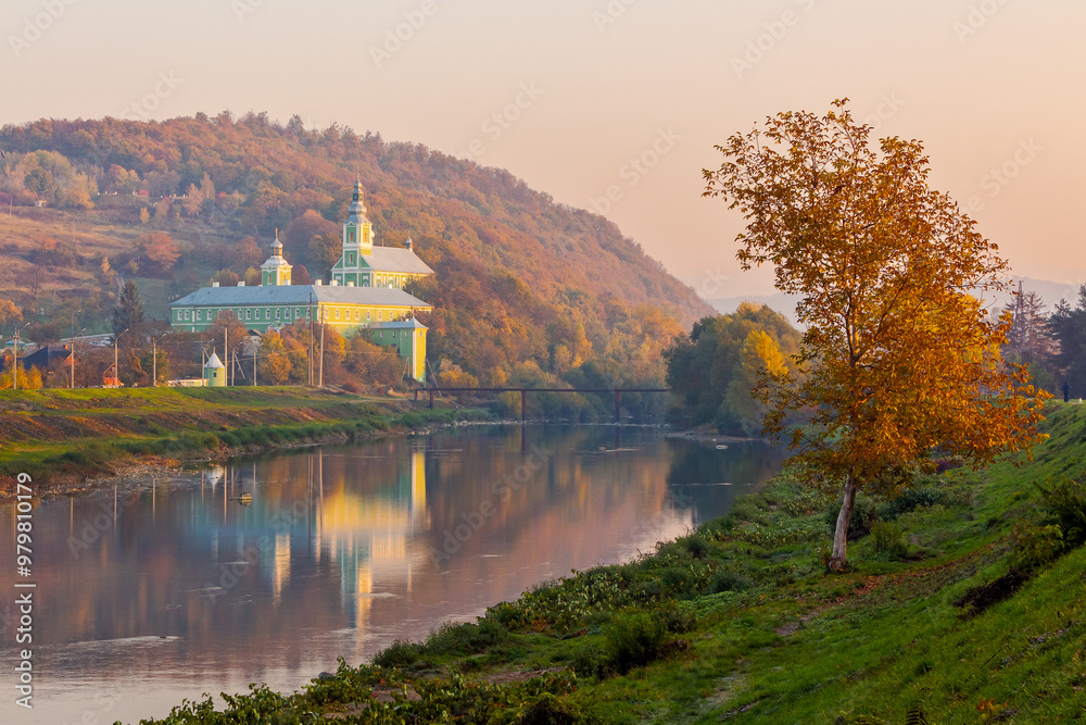Fototapeta premium mukachevo, ukraine - 06 nov 2015: old town with latorica river in autumn. embankment with trees in fall season. saint nicholas monastery in the distance. urban scenery in morning light