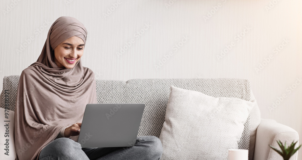 Modern arab girl in hijab using laptop at home, working remotely while sitting on couch in living room Modern arab girl in hijab using laptop at home, working remotely while sitting on couch in living room