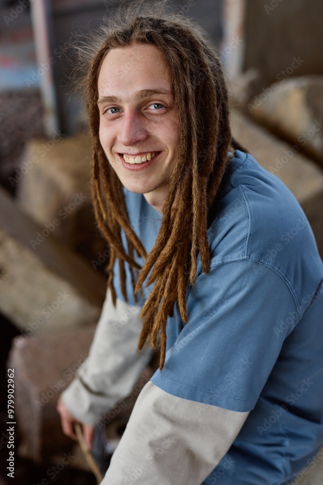 Young man with dreadlocks smiling while sitting on rocky surface and wearing a blue shirt and light jacket, creating an informal and relaxed environment Young man with dreadlocks smiling while sitting on rocky surface and wearing a blue shirt and light jacket, creating an informal and relaxed environment