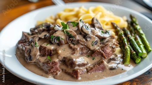 Beef stroganoff with tender beef slices and creamy mushroom sauce, served with a side of buttered egg noodles and roasted asparagus, on a white plate photo