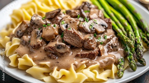 Beef stroganoff with tender beef slices and creamy mushroom sauce, served with a side of buttered egg noodles and roasted asparagus, on a white plate photo