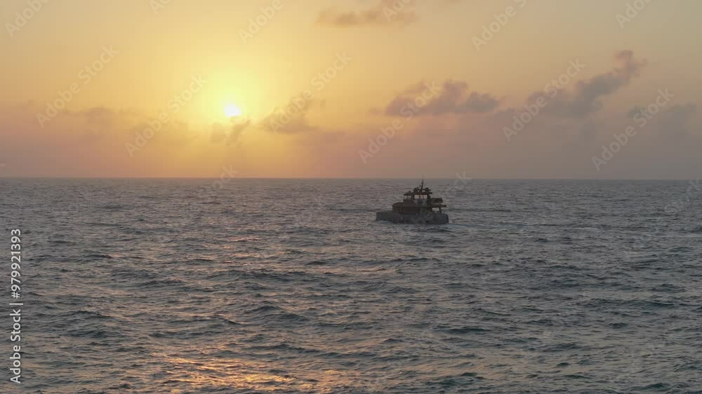 A motorboat heads toward the sunrise, filmed in slow motion by a drone. The calm sea reflects the orange sun just above the horizon, with scattered clouds overhead.
