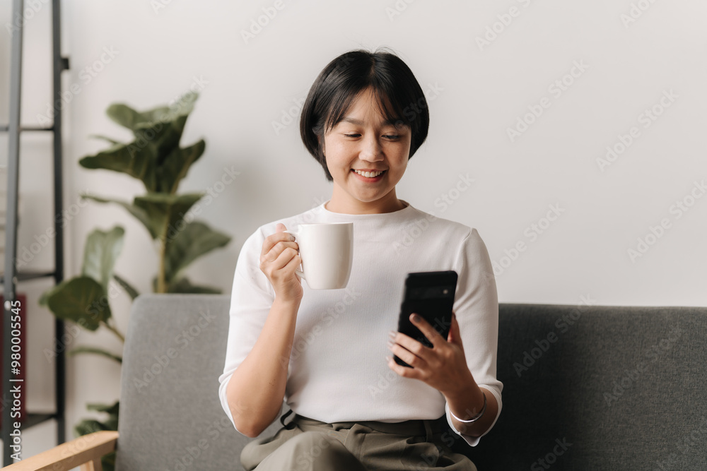 Morning Coffee and Connection: A young Asian woman enjoys her morning routine, checking her phone with a warm beverage in hand, capturing the essence of modern relaxation and connectivity. Morning Coffee and Connection: A young Asian woman enjoys her morning routine, checking her phone with a warm beverage in hand, capturing the essence of modern relaxation and connectivity.
