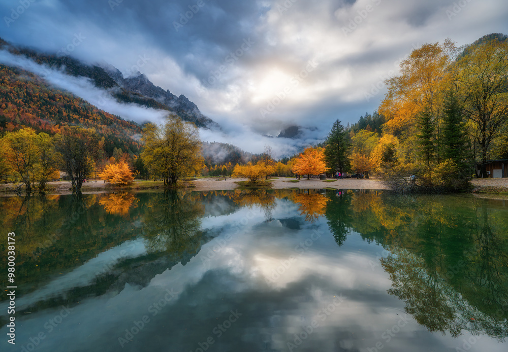 Mountain lake reflecting colorful trees with red and orange foliage at autumn sunset. Dramatic landscape with water, foggy forest, mountains in low clouds in fall. Jasna lake, Kranjska Gora, Slovenia Mountain lake reflecting colorful trees with red and orange foliage at autumn sunset. Dramatic landscape with water, foggy forest, mountains in low clouds in fall. Jasna lake, Kranjska Gora, Slovenia