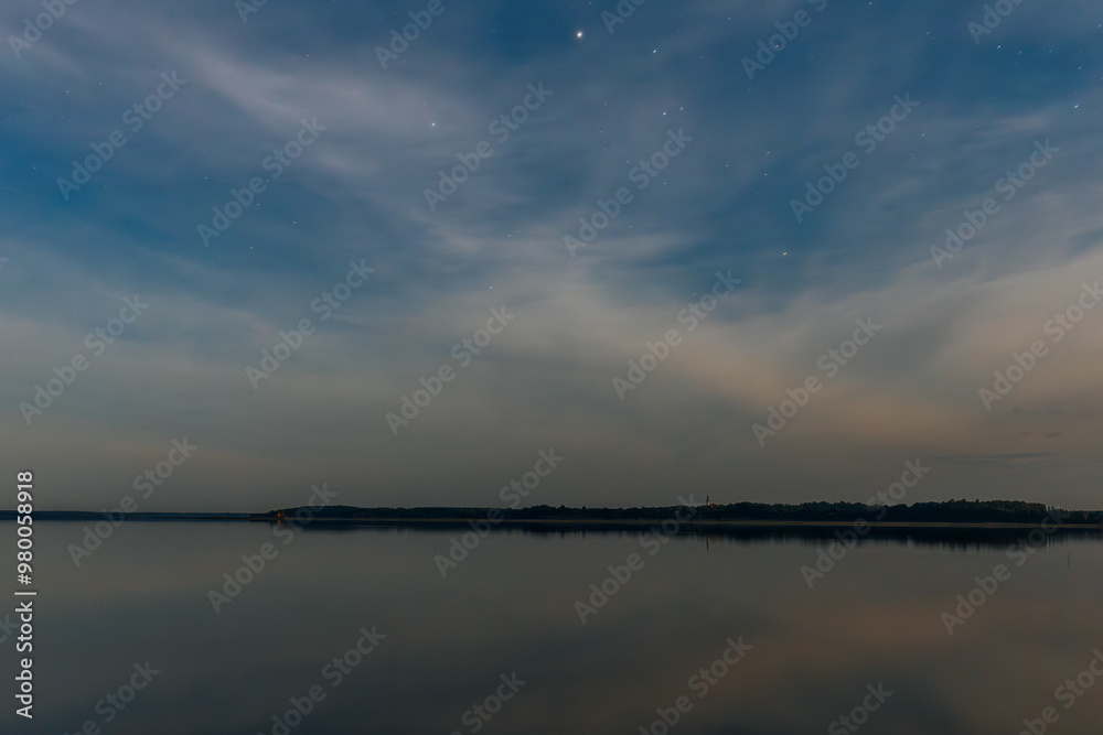 A tranquil lake at dusk, with small boats lined along the shore. The smooth water reflects the evening sky, filled with soft clouds and a few stars. The calm atmosphere creates a peaceful