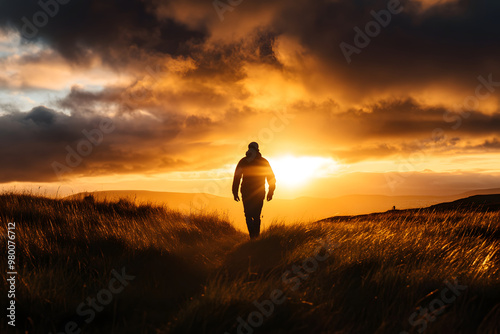 A lone individual strolls through a lush grassy area during sunset, as vibrant golden sunlight casts long shadows and highlights the clouds above. Take a hike day photo