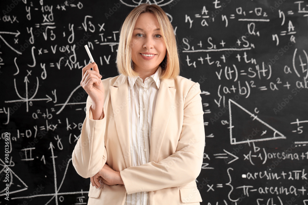 Mature female math teacher with chalk near blackboard in classroom Mature female math teacher with chalk near blackboard in classroom