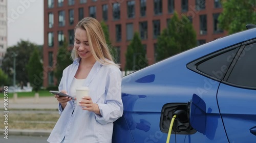 Pretty woman with disposable coffee cup and cellphone checks electric car charging on street. Young long-haired blonde highlights modern eco-friendly lifestyle