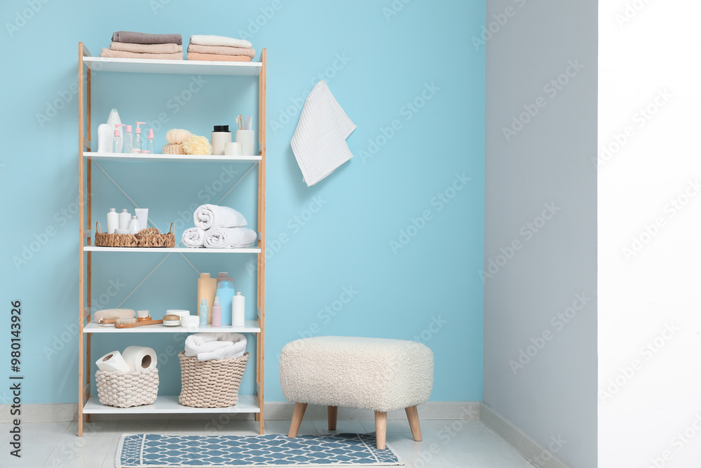 Fototapeta premium Modern shelving unit with bath accessories and pouf near blue wall in room