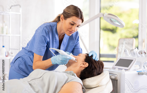 Skilled professional female aesthetician performing radiofrequency facial skin tightening procedure on mature woman in modern cosmetology clinic..Skilled professional female aesthetician performing