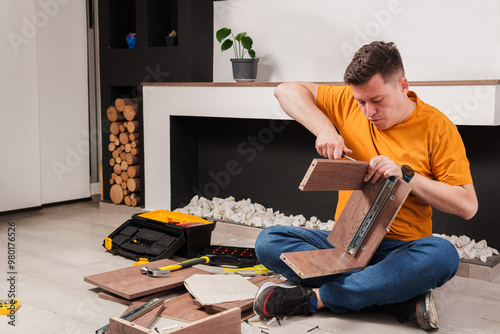 A young hard-working man is assembling a piece of furniture on the living room floor of an apartment. He has a screwdriver in his hand and is putting together some parts.