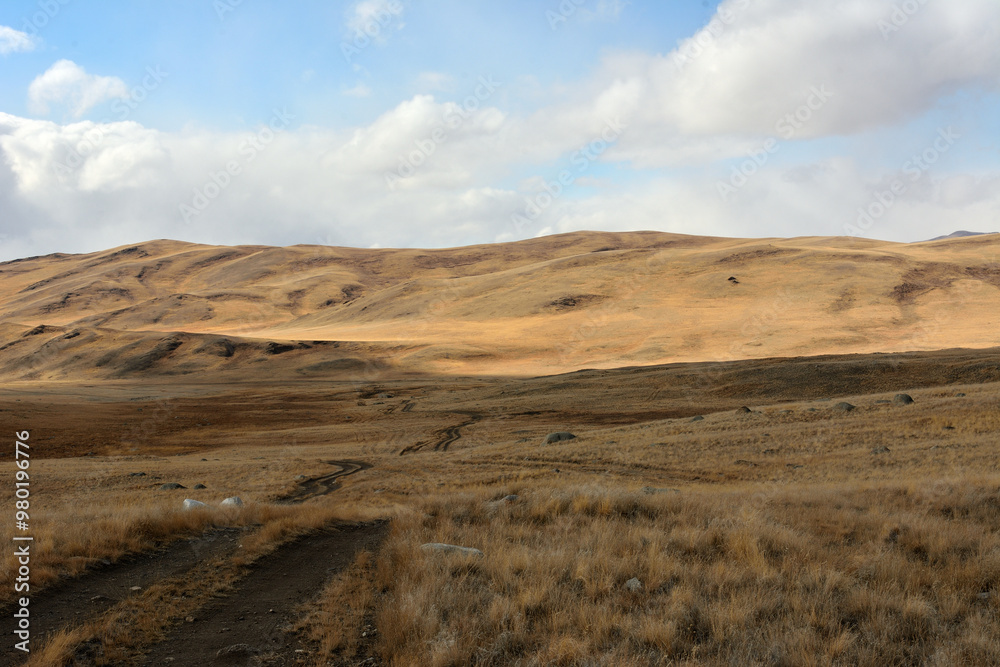A muddy field road descends into a hilly steppe with dry, yellowed grass on an autumn sunny day. A muddy field road descends into a hilly steppe with dry, yellowed grass on an autumn sunny day.