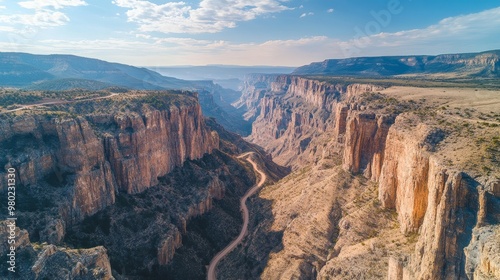 Aerial view of a grand canyon with jagged rock formations, deep gorges, and a winding trail cutting through the rugged landscape under a bright blue sky