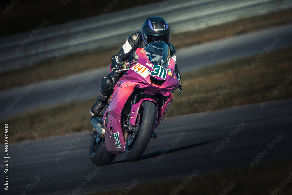 A person rides a pink motorcycle on a race track, surrounded by lush grass and a fence in the background A person rides a pink motorcycle on a race track, surrounded by lush grass and a fence in the background