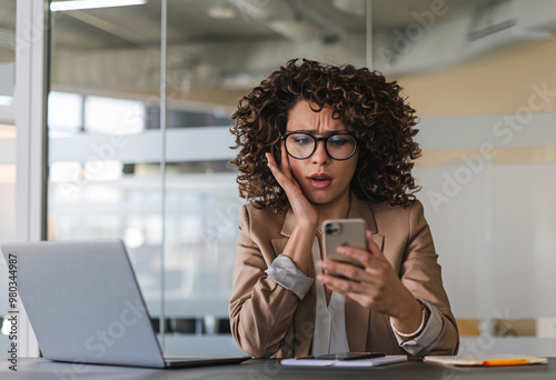 Frightened young African American woman sitting at her office desk, looking at her mobile phone screen in disbelief, holding her hand to her cheek.