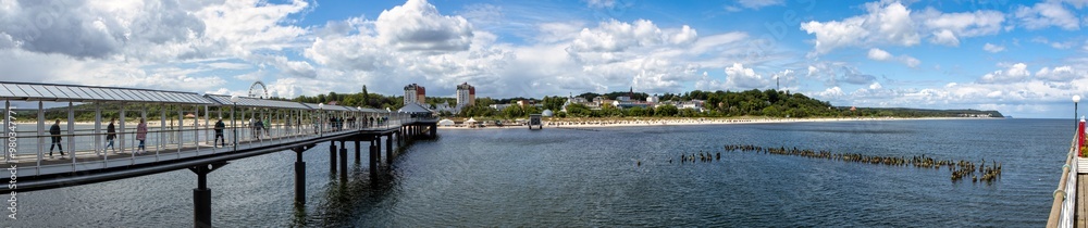 Obraz premium Stunning Panorama of a Coastal Landscape Featuring a Sandy Beach and Charming Small Town Viewed from the Pier, Under a Clear Blue Sky with Fluffy Clouds, Perfect for Summer Vibes