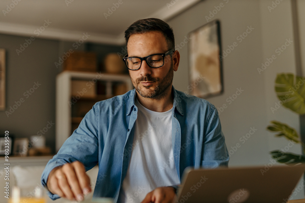 Fototapeta premium A focused mid aged male freelancer sitting at the wooden table at home and reading online projects on a laptop.