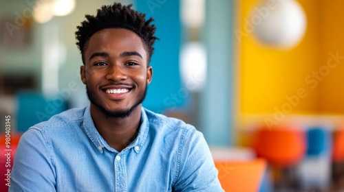 A confident young African American man sits in a lively office environment, smiling broadly while wearing a blue shirt. Bright colors and contemporary design enhance the atmosphere photo