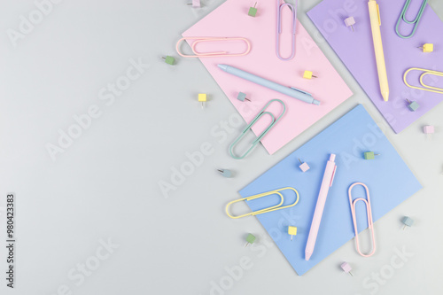 Top view of pink, purple and blue notebooks on grey background. Colorful pens, paperclips, pins, school, office wallpaper. Flat lay, copy space.