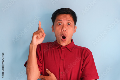 Asian young man shocked in red shirt is pointing his finger to the up at an empty space.