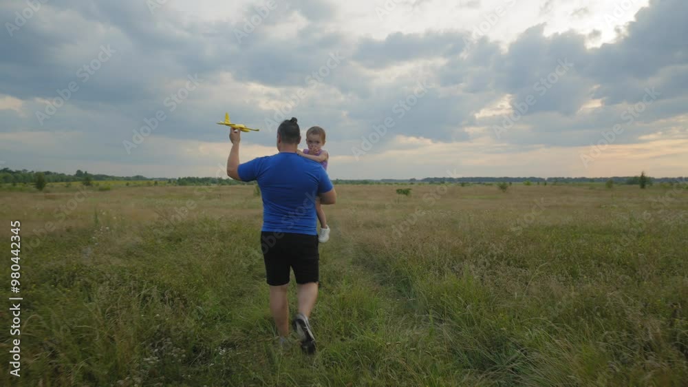 Rear view of handsome father carrying cute toddler baby daughter, walking across green field and playing with toy airplane , imagining to fly while happy family relaxing at countryside at sunset. Rear view of handsome father carrying cute toddler baby daughter, walking across green field and playing with toy airplane , imagining to fly while happy family relaxing at countryside at sunset.