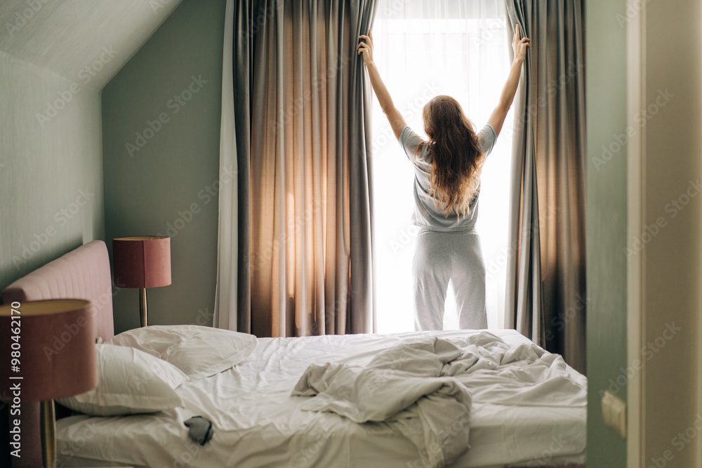 Woman opening curtains in bedroom and looking out.