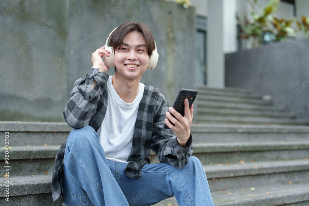 Obraz premium Happy young Asian student wearing wireless headphones and smiling happily while looking at mobile phone at university, college, field trip Learning concept, success.