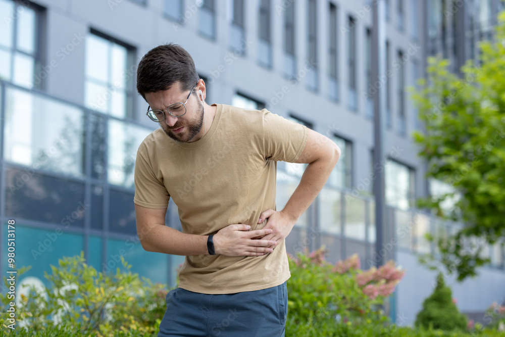 Tired and grimacing young man standing outside on the street and holding the side of his stomach after a workout and a run Tired and grimacing young man standing outside on the street and holding the side of his stomach after a workout and a run