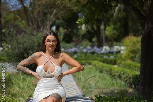 A young, dark-haired, beautiful woman, heavily tattooed, wearing a white dress and sunglasses with purple lenses, sitting, with her hands on her waist, looking at the camera relaxed and calm.