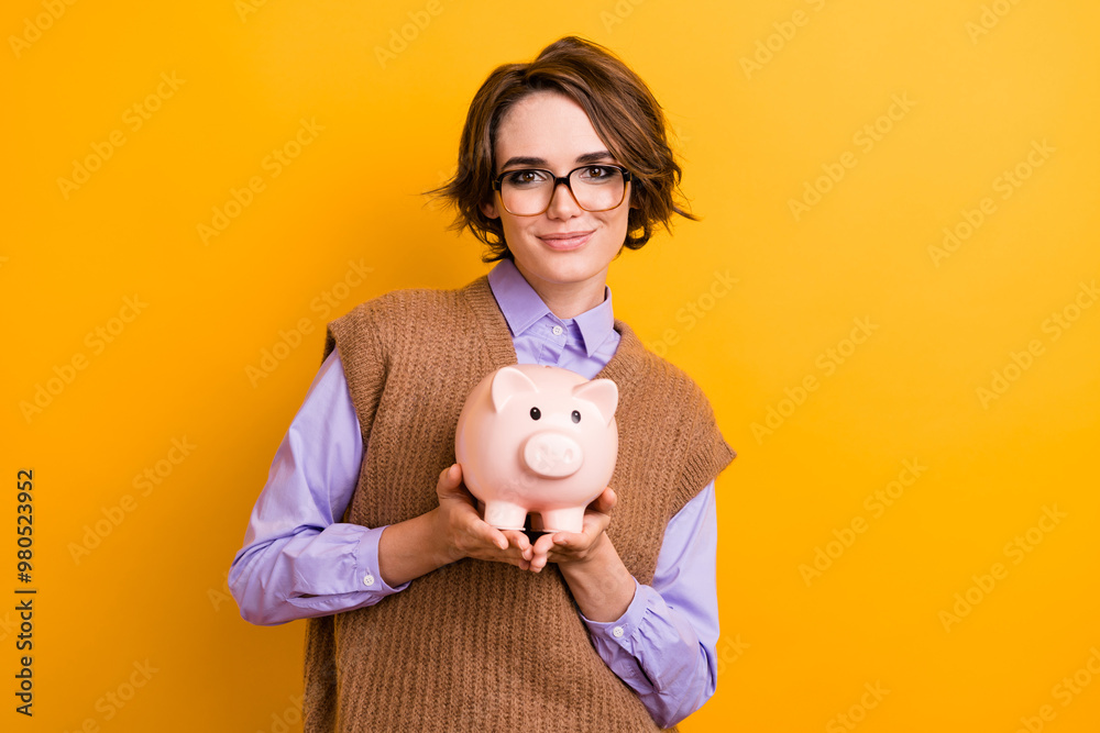 Photo of good mood positive girl dressed knitwear waistcoat in glasses hold money piggy box in arms isolated on yellow color background Photo of good mood positive girl dressed knitwear waistcoat in glasses hold money piggy box in arms isolated on yellow color background