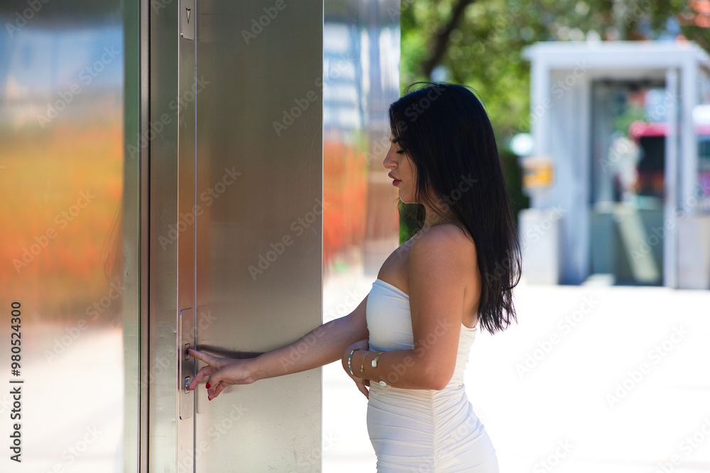Young, brunette, beautiful Spanish woman in a white dress pressing the call button of a steel and glass lift. The woman goes down to the car park to pick up her car. Young, brunette, beautiful Spanish woman in a white dress pressing the call button of a steel and glass lift. The woman goes down to the car park to pick up her car.